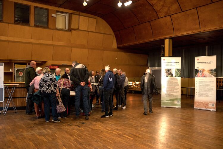 Ca. 20 Personen stehen in einer holzverkleideten Halle um einen Tisch, rechts zwei Roll-ups zum Projekt