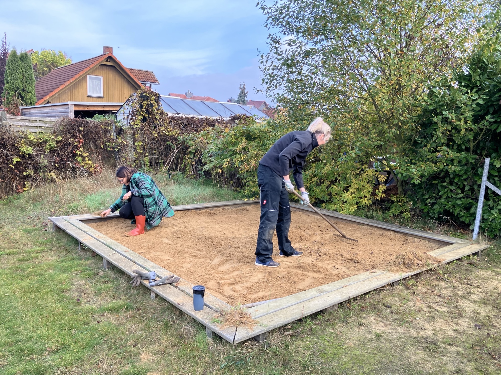 Großer Sandkasten in einem Garten, eine Frau kniet im Sand, eine Frau recht den Sand