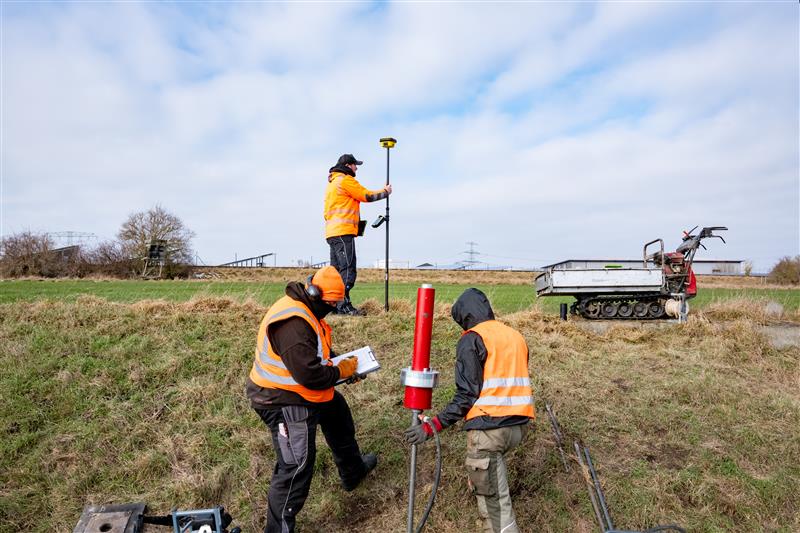 Drei Bauarbeiter stehen mit Bohrer und Erkundungswerkzeugen auf der Wiese.